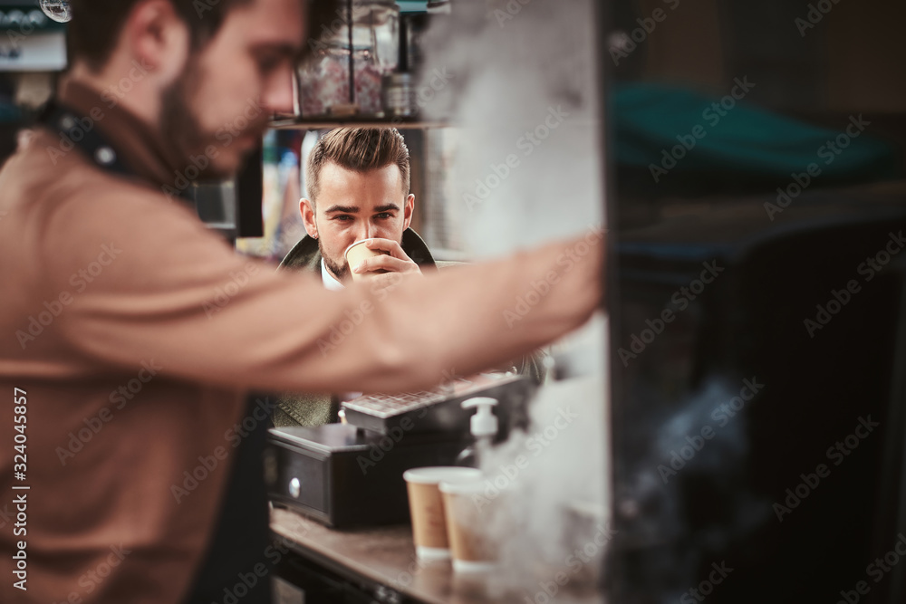 Hipster and elegant young male customer sitting outdoors next to coffee making barista in a mobile coffee shop in a city emporium, wearing green wool coat, white shirt and bow tie, taking a sip of