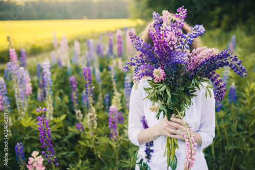 Fototapeta Naklejka Na Ścianę i Meble -  Beautiful woman in a white dress hid her face behind a large bouquet of lupins