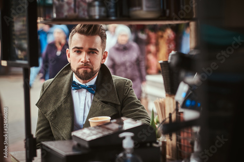 Stylish and elegant young male customer sitting outdoors next to coffee making gear in a mobile coffee shop in a city emporium, wearing green wool coat, white shirt and bow tie, holding a paper cup of