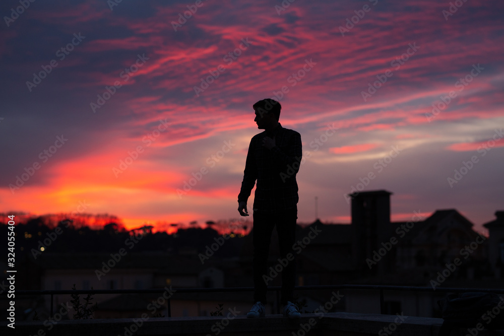 Black silhouette of man on sunset above the orange and rose clouds. Young man standing on top of a hill. Concept of freedom, happiness, success, victory. Guy looking at gorgeous sunset in city. Black silhouette of man on sunset above the orange and rose clouds. Young man standing on top of a hill. Concept of freedom, happiness, success, victory. Guy looking at gorgeous sunset in city.