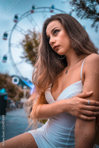 Street style, portrait of a young brunette in white dress in front of the tourist ferris wheel in the city