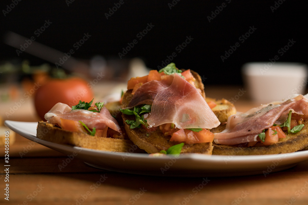 Bruschetta with tomato, basil, garlic and Italian ham on a white plate surrounded by ingredients, warm light closeup Bruschetta with tomato, basil, garlic and Italian ham on a white plate surrounded by ingredients, warm light closeup