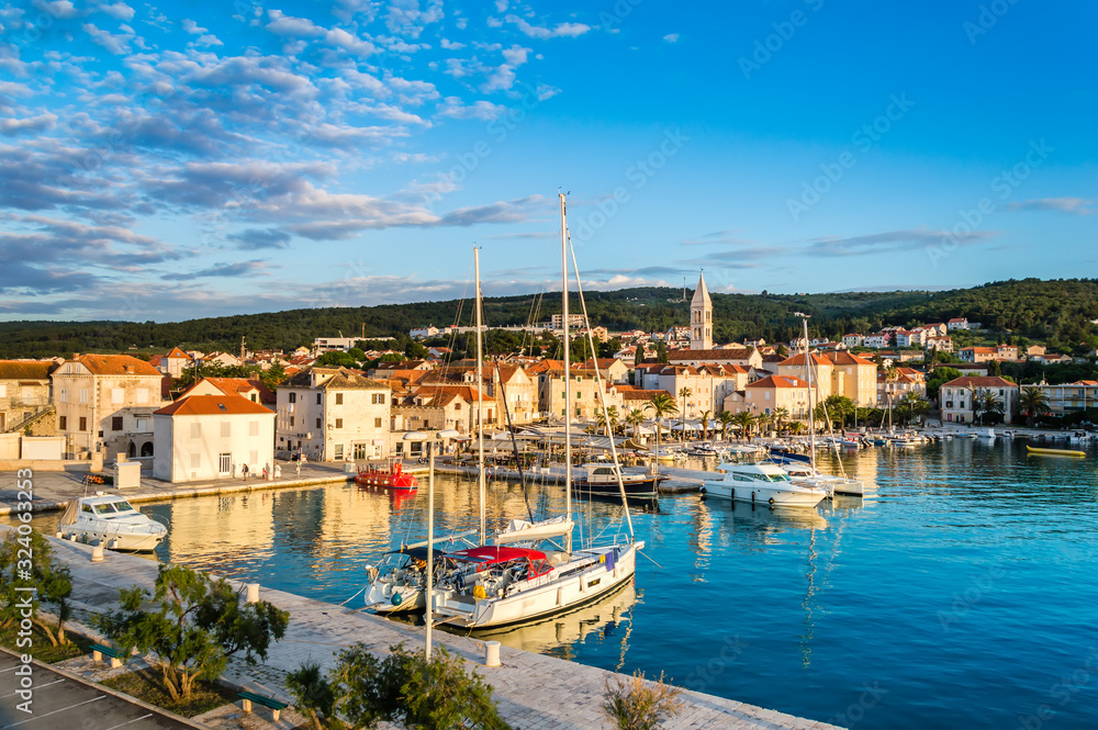 Supetar on Brac Island near Split, Croatia. Small seaside town with promenade and harbor with white boats, palm trees, cafes, houses and church. Tourists walk the street on sunny day at sunset