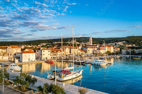 Supetar on Brac Island near Split, Croatia. Small seaside town with promenade and harbor with white boats, palm trees, cafes, houses and church. Tourists walk the street on sunny day at sunset photo