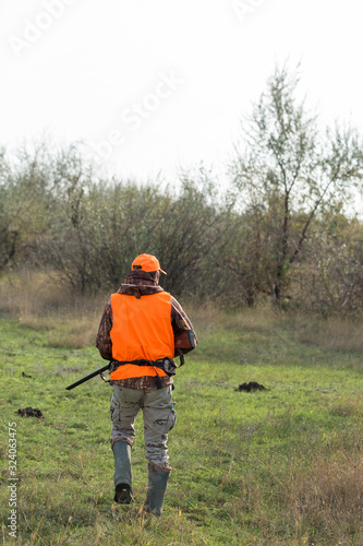 A man with a gun in his hands and an orange vest on a pheasant hunt in a wooded area in cloudy weather. Hunter with dogs in search of game.