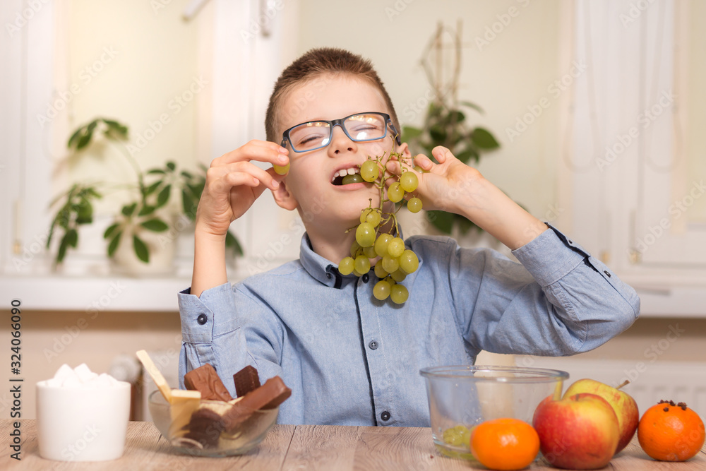 Uśmiechnięty chłopiec siedzi przy stole i zjada owoce. Trzyma w zębach owoce winogrona.