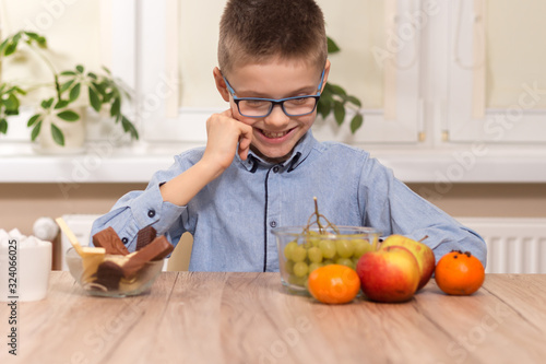 Chłopiec wieku szkolnym siedzi przy stole i z uśmiechem wpatruje się w owoce leżące na stole. Słodycze stojące na stole chłopiec ignoruje.