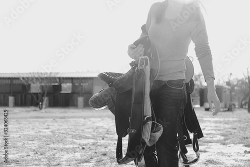 Western lifestyle shows woman carrying saddle close up in black and white.  Vintage style farm and ranch scene.