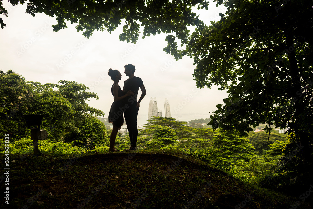 Silhouette of young couple standing underneath trees in a forest, skyscrapers in the distance.,Singapore Silhouette of young couple standing underneath trees in a forest, skyscrapers in the distance.,Singapore