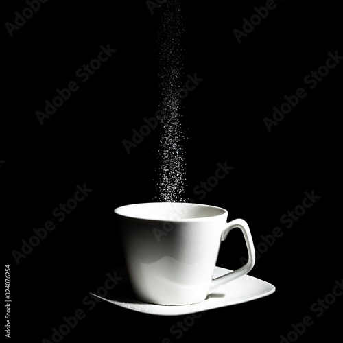A low key photography of a white cup on top of a saucer, getting sugar poured into it in front of a complete black background