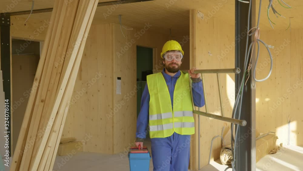 SLOW MOTION, CLOSE UP: Worker coming to work carries his metal toolbox and ladder across the room of a prefabricated house. Builder leaves the construction site with his toolbox and ladder in hand. SLOW MOTION, CLOSE UP: Worker coming to work carries his metal toolbox and ladder across the room of a prefabricated house. Builder leaves the construction site with his toolbox and ladder in hand.