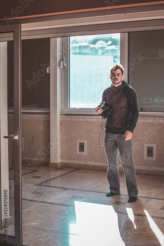 Man standing in an apartment next to a window holding a vintage photo camera in his hand. Copyspace on left. © Anze