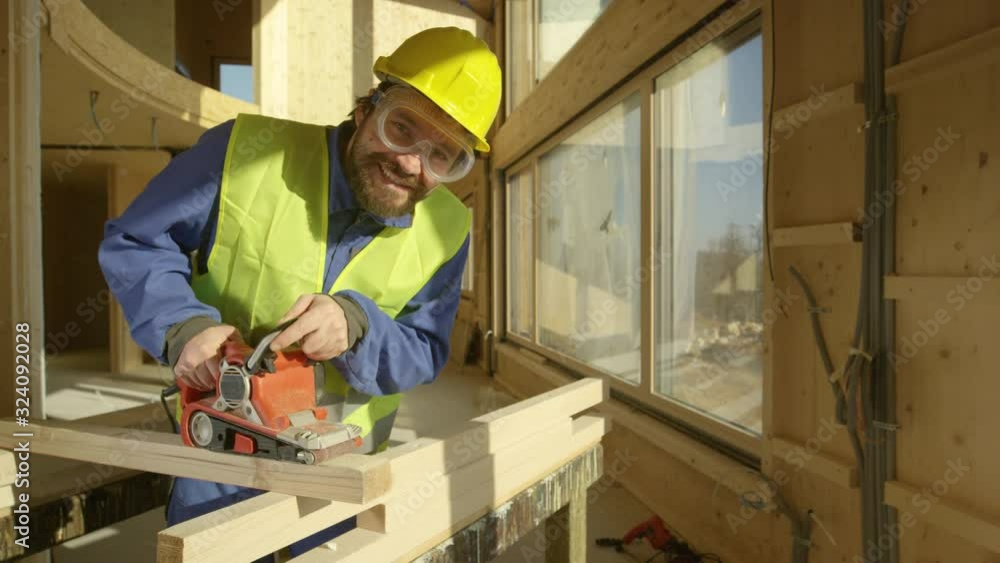 SLOW MOTION, PORTRAIT, CLOSE UP: Smiling builder working on a prefabricated house buffs a long wooden beam. Cheerful Caucasian contractor uses an electric sander to smooth out the edge of a plank. SLOW MOTION, PORTRAIT, CLOSE UP: Smiling builder working on a prefabricated house buffs a long wooden beam. Cheerful Caucasian contractor uses an electric sander to smooth out the edge of a plank.