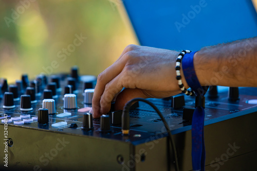 A close up view on the hand of an electronic music DJ turning dials on a CDJ mixer, wearing bangles against green blurry background at earth festival