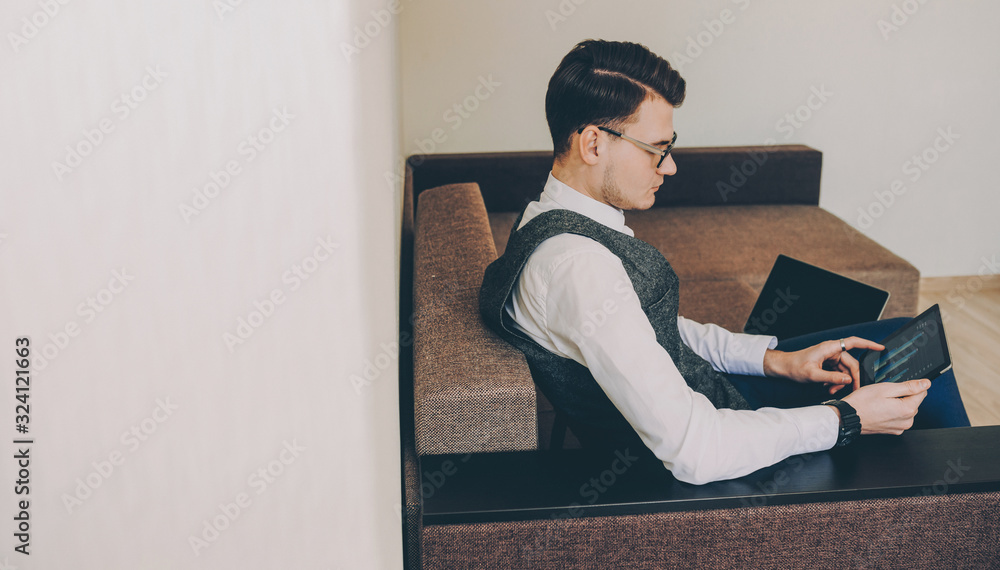 Lateral view of a confident caucasian freelancer with eyeglasses working on his tablet sitting on a sofa