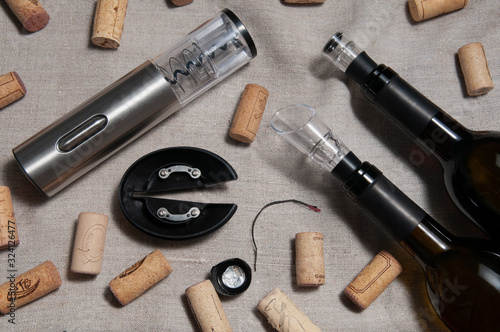 Electric corkscrew in steel gray. Lies on a linen background. Nearby are several wine corks and two bottles of wine.