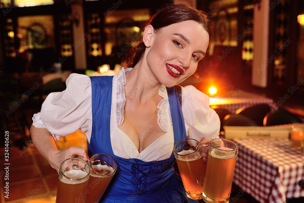 an attractive young girl in Bavarian clothes holds a lot of mugs with beer on the pub background during the celebration of OKtobfest