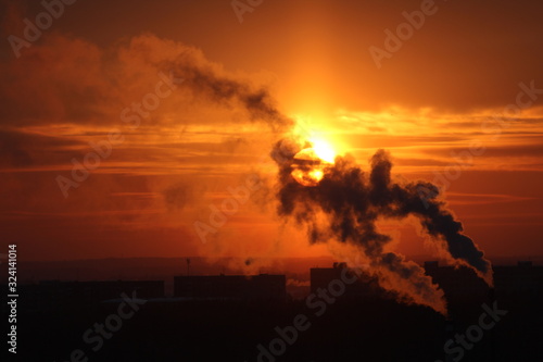 Red sun at sunset or dawn at dusk low above the horizon shrouded in smoke from the chimneys of the plant