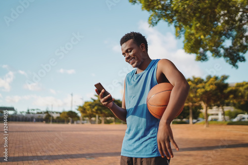 Fototapeta Naklejka Na Ścianę i Meble -  Happy young ,ale sportive basketball player holding ball in his arms using mobile phone at park