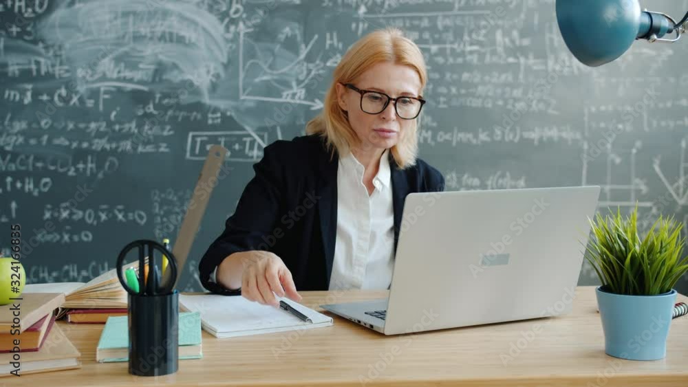University professor serious adult lady in suit and glasses is using laptop then taking notes working at desk in class. Technology and education concept. University professor serious adult lady in suit and glasses is using laptop then taking notes working at desk in class. Technology and education concept.