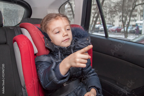 girl in blue warm clothes sits unbuckled in a car seat and points her hand forward photo