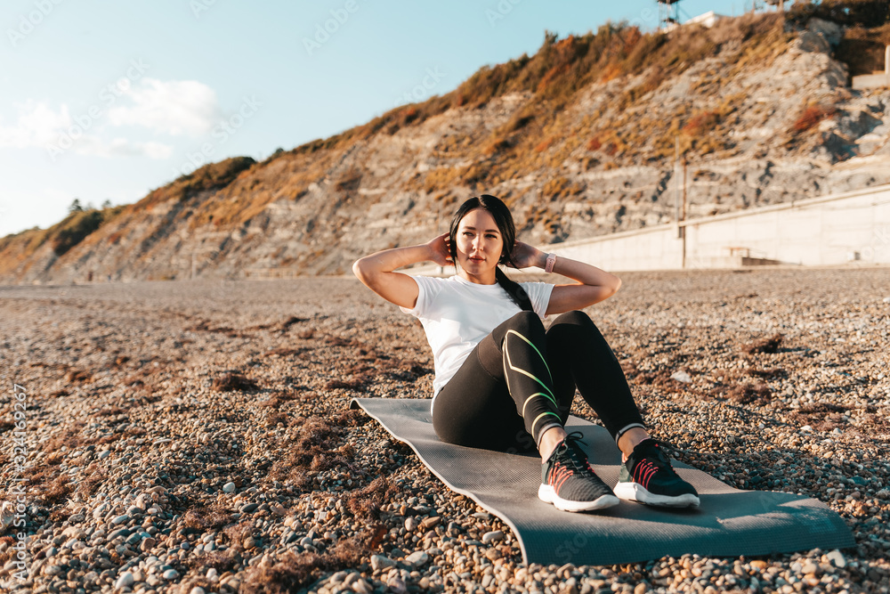 The concept of sport. A young brunette woman shakes her abs sitting on a sports Mat. In the background mountains and wild beach The concept of sport. A young brunette woman shakes her abs sitting on a sports Mat. In the background mountains and wild beach
