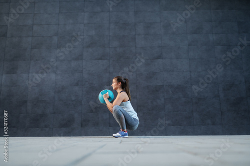 Side view of caucasian sportswoman in shape holding weight ball while crouching and doing endurance. In background is dark wall. © dusanpetkovic1