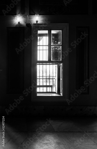 Black and white high contrast shot of narrow window, revealing strong light into dark room with illuminated lanterns and white tiled marble floor