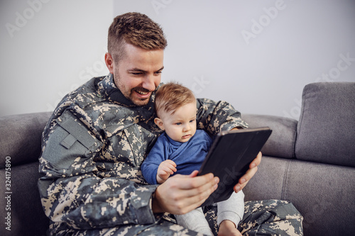Young bearded soldier sitting on sofa in living room with his beloved son and they are watching cartoons on tablet.
