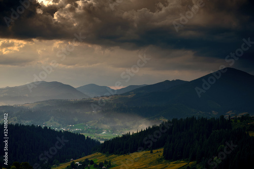 Golden sunset in carpathian mountains - beautiful summer landscape, spruces on hills, village, homes, dark cloudy sky and bright sun light, meadow and wildflowers