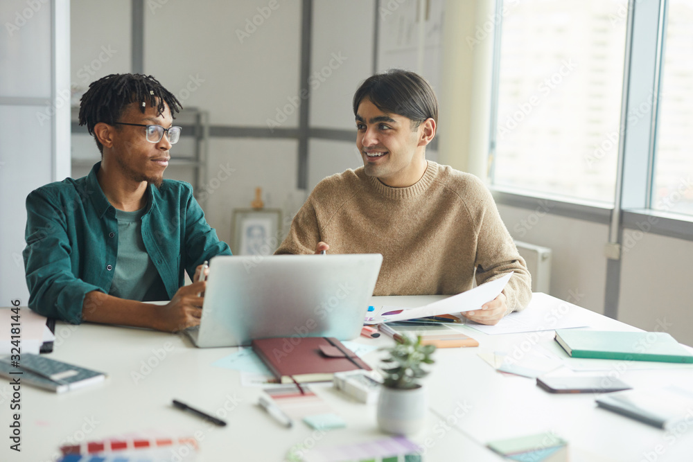 Two young businessmen in casual clothing sitting at the table talking and using laptop in work at office Two young businessmen in casual clothing sitting at the table talking and using laptop in work at office