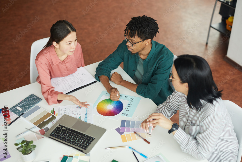Multiethnic group of graphic designers sitting at the table with colored palette and choosing the color during their work at office Multiethnic group of graphic designers sitting at the table with colored palette and choosing the color during their work at office