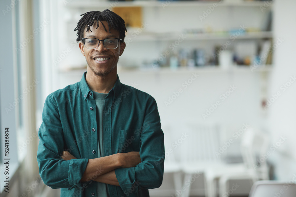 Obraz premium Portrait of African businessman in eyeglasses standing with arms crossed and smiling at camera at office