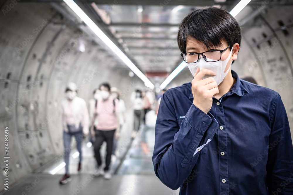 Asian man wearing surgical face mask coughing in subway station with crowded people walking pass. Wuhan coronavirus (COVID-19) outbreak prevention in public area. Health care and medical concept Asian man wearing surgical face mask coughing in subway station with crowded people walking pass. Wuhan coronavirus (COVID-19) outbreak prevention in public area. Health care and medical concept