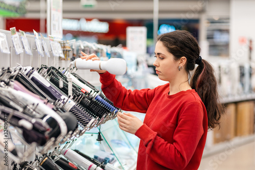 Caucasian young woman with long hair  chooses a hair dryer. In the background  shelves with household appliances. The concept of buying household appliances for beauty and self-care