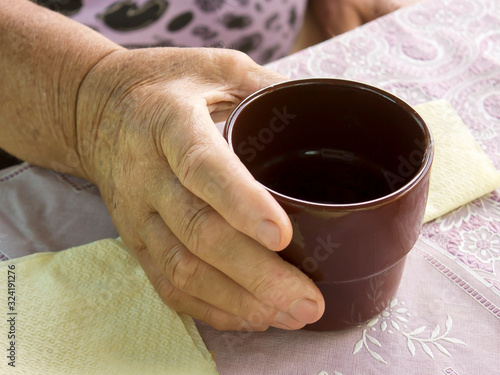 Hand of an old woman holding a mug. A woman is sitting at the table.