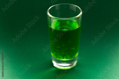 A green drink in a transparent glass stands on a green background. Carbonated liquid in a shot glass. Drink in a glass