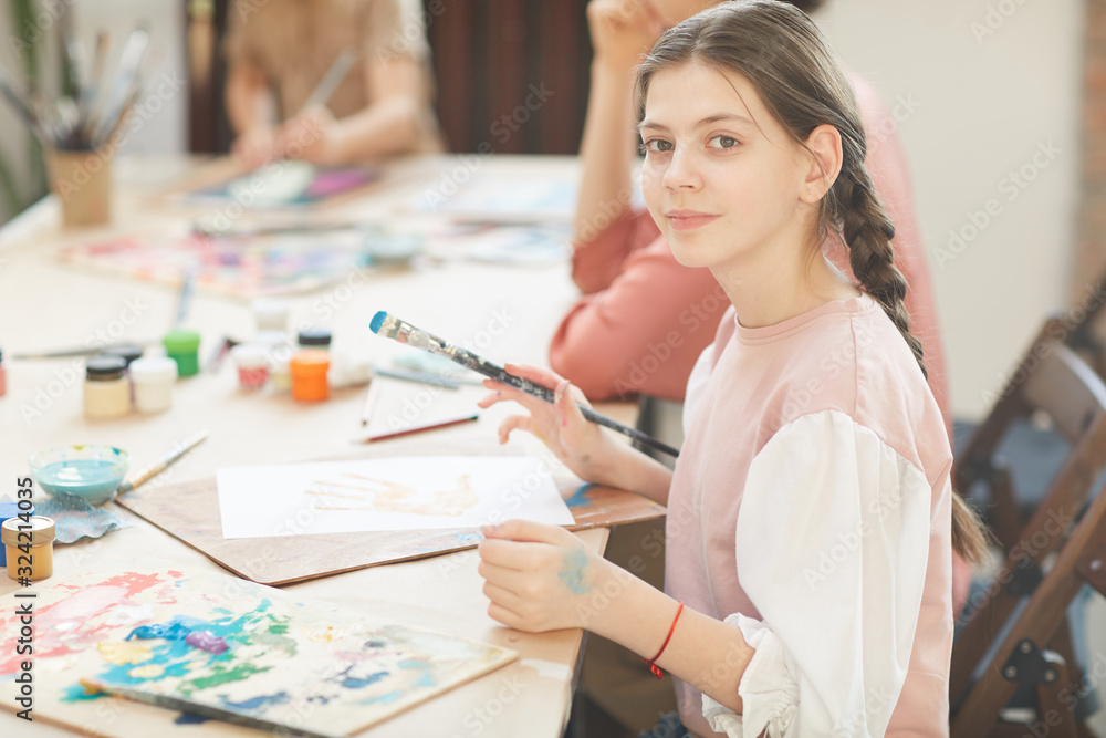 Portrait of girl looking at camera while sitting at the table with papers and paints and painting during art lesson