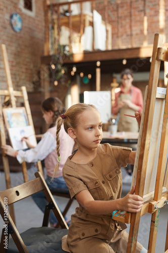 Little girl sitting in front of easel and painting with other students during art lesson at art studio
