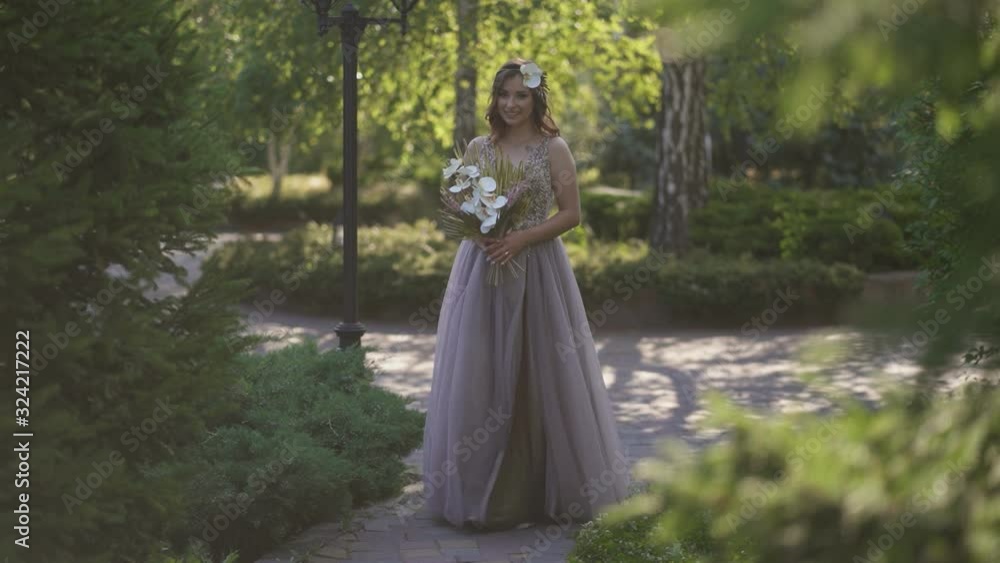 A young beautiful happy girl, a bride with a beautiful bouquet in her hands walks through the park, dressed in a colorful wedding dress, looks at the sunlight and the camera lens A young beautiful happy girl, a bride with a beautiful bouquet in her hands walks through the park, dressed in a colorful wedding dress, looks at the sunlight and the camera lens