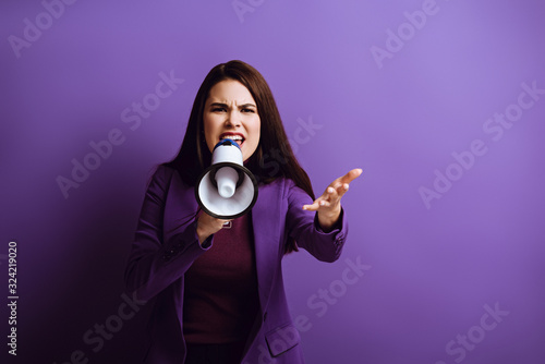 irritated young woman screaming in megaphone while showing indignation gesture on purple background