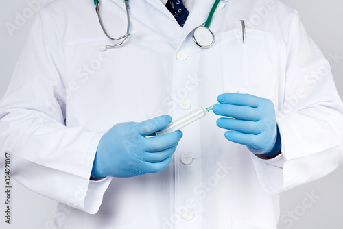 Fototapeta Naklejka Na Ścianę i Meble -  male doctor in a white coat and tie stands and holds a plastic syringe with a needle on a white background, wearing blue sterile medical gloves
