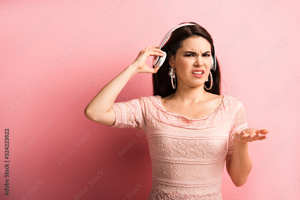 Fototapeta premium displeased girl touching wireless headphones while standing with open arm on pink background
