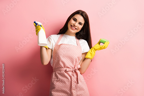 happy housewife in apron and rubber gloves holding spray bottle and sponge on pink background photo
