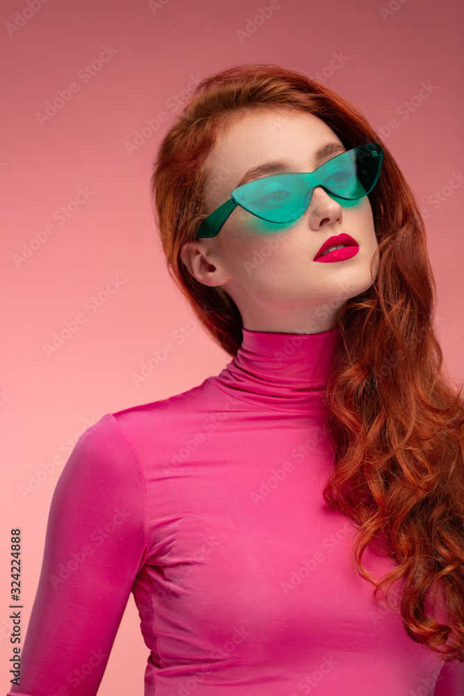 Close-up portrait of smiling redhead girl. Perfect skin. Beautiful female model with long hair over pink background. Young cute woman in green sunglasses with clear skin. Close-up portrait of smiling redhead girl. Perfect skin. Beautiful female model with long hair over pink background. Young cute woman in green sunglasses with clear skin.