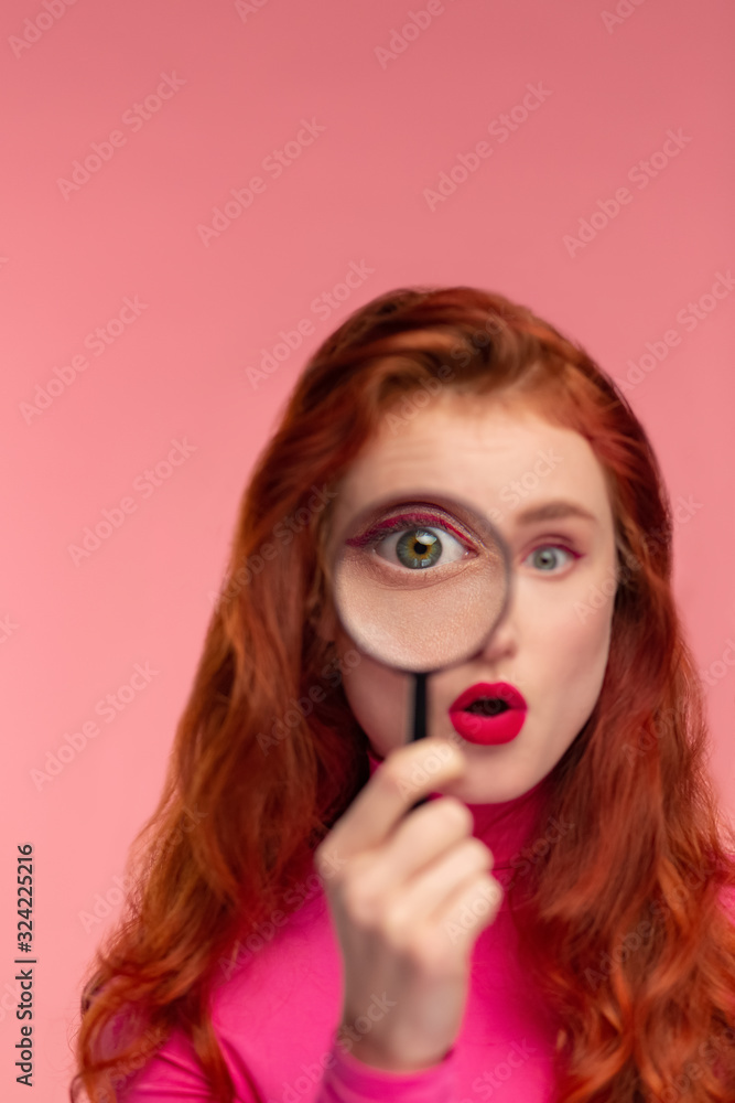 Young detective. Close-up portrait of surprised redhead girl looking at camera through magnifying glass, over pink background. Funny model with long hair starting investigation. Woman's intuition.