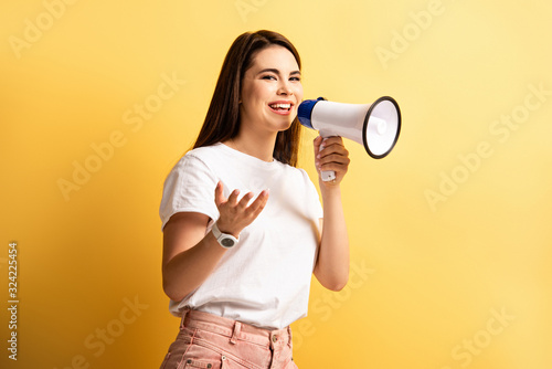 cheerful girl speaking in megaphone while standing with open arm on yellow background