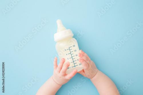 Infant hands holding  bottle of milk on light blue floor background. Feeding time. Pastel color. Closeup. Point of view shot.