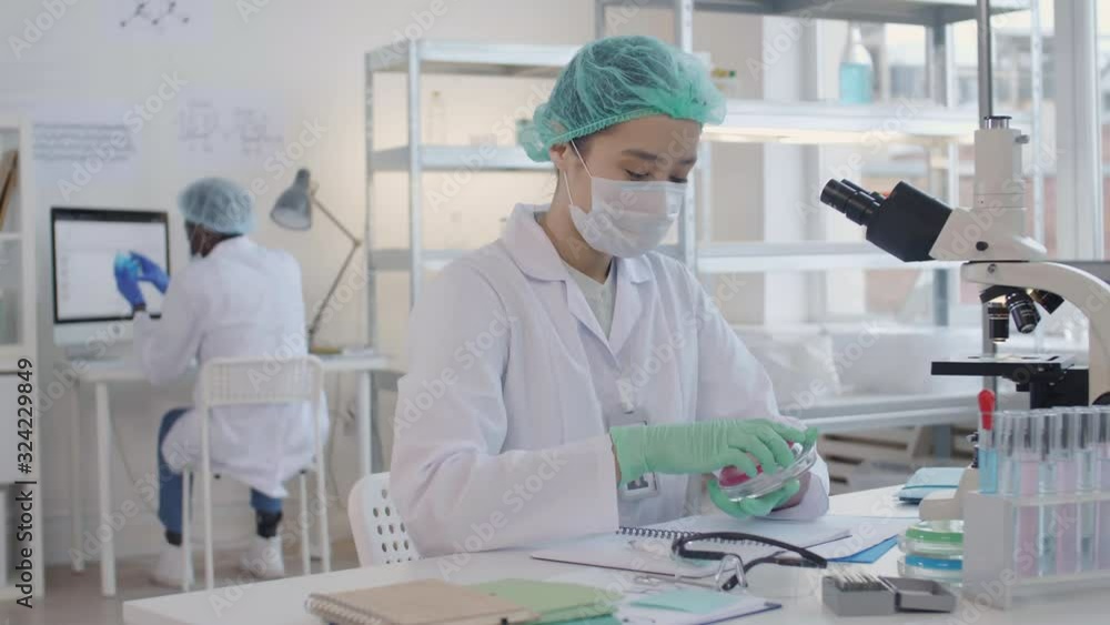 Lockdown of female Asian scientist wearing medical overall, protective hat, mask and gloves examining petri dish and making notes in notebook Lockdown of female Asian scientist wearing medical overall, protective hat, mask and gloves examining petri dish and making notes in notebook