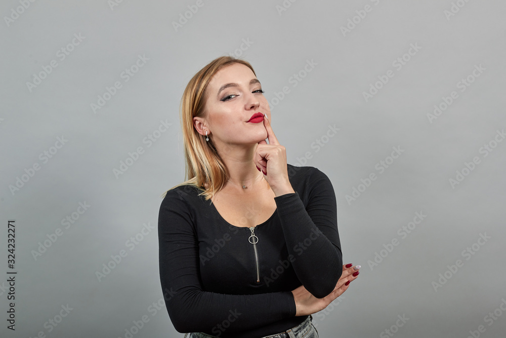 Young blonde girl in black jacket on grey background sly woman thinks, holds her chin on hand Young blonde girl in black jacket on grey background sly woman thinks, holds her chin on hand